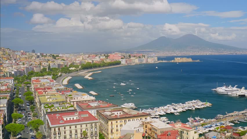 Naples, Italy. Scenic aerial view of the Gulf with elegant marina, yachts, waterfront promenade, colorful city buildings, and Mount Vesuvius rising in the distance under a clear sky. 4K pan video.