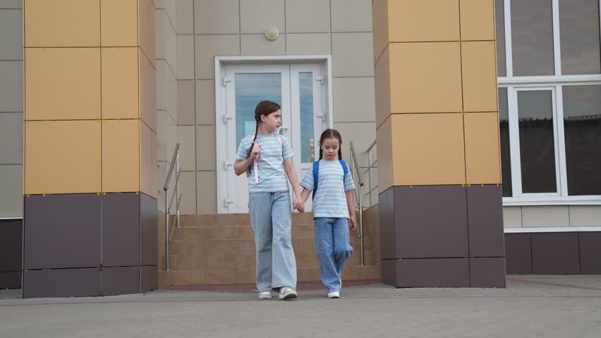 Two schoolgirls child walk along sidewalk, one them holds other by hand. child kid carry backpacks walk building. happy family. girl child kid. child walk happy family children dream. happy family