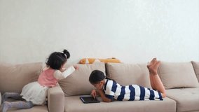 Asian sisters at home, sitting together on beige sofa. Older girl lies relaxed, scrolling on tablet, while sister shows impatience and demands attention. Sibling relationship, childhood emotions. - Powered by Shutterstock - Get 15% off with code: PIKWIZARD15