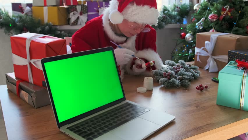 Woman in festive costume paints a red and white candy cane decoration by hand while sitting at a wooden table with wrapped gifts and holiday wreath. A laptop with green screen is placed nearby.