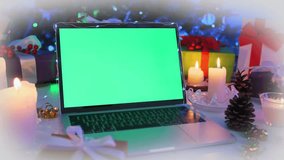 Laptop computer with green chroma key screen standing on decorated table with candles, wrapped gifts, pine cones, and festive Christmas tree lights in background. - Powered by Shutterstock - Get 15% off with code: PIKWIZARD15