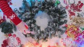 Close-up of a person in a festive sweater spraying artificial snow on a green Christmas wreath on a white table with red ornaments and golden ribbons, preparing for holiday home decoration. - Powered by Shutterstock - Get 15% off with code: PIKWIZARD15