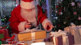 Bearded Santa Claus carefully wraps a golden Christmas gift box, cutting ribbon with scissors at festive table. Cozy holiday atmosphere with Christmas tree, sparkling garlands, and bright decorations. - Powered by Shutterstock - Get 15% off with code: PIKWIZARD15