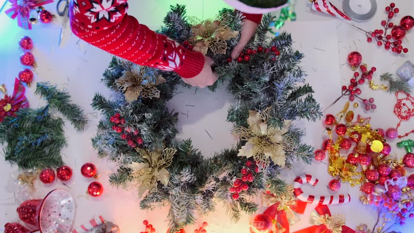 Top view of hands decorating a Christmas wreath with golden flowers and red berries, surrounded by festive ornaments and holiday decor. - Powered by Shutterstock - Get 15% off with code: PIKWIZARD15