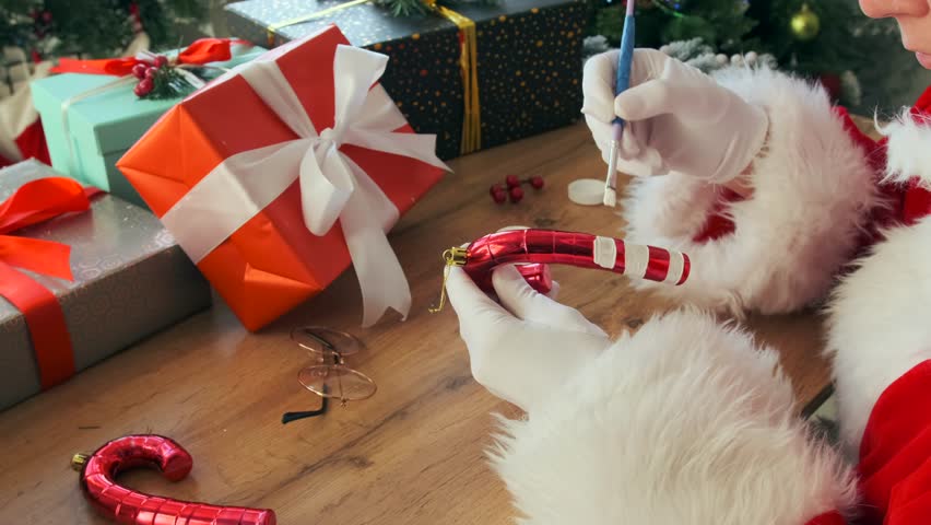 Close-up of Santa Claus in white gloves carefully painting a red and white candy cane Christmas ornament with a brush at a decorated wooden table. Brightly wrapped presents and festive decorations.