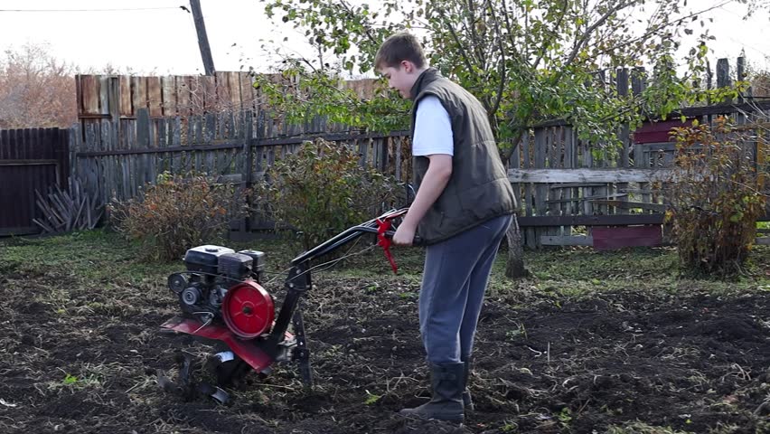 Young boy with light brown hair operates a tiller in a garden, wearing a green vest and gray pants, showcasing agricultural work and outdoor activity in a rural setting	