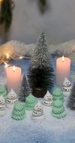 Sweet meringue christmas cookies shaped like snowmen and fir trees creating a magical winter scene with flickering candles, artificial snow, and festive holiday decorations on a blue surface