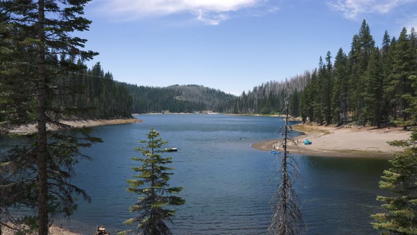 Wide view of a lake surrounded by dense pine forest. On the sandy shore to the right, there is a blue tent with kayaks nearby. In the center of the scene, calm water with a floating dock can be seen