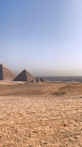 A vertical shot of the Great Pyramids of Giza rising from the sandy desert, with a hazy cityscape visible in the distance under a clear blue sky. 4k ultra hd video