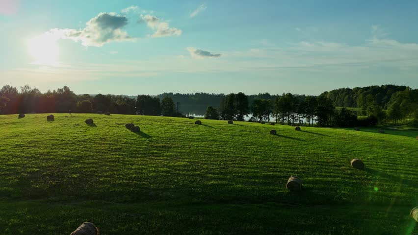 Golden sunlight shines on a vibrant green field with scattered hay bales under a blue sky with soft clouds.