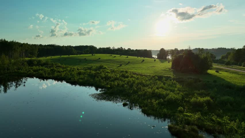 Green field of hay bales under a partly cloudy sky in the late afternoon sun.
