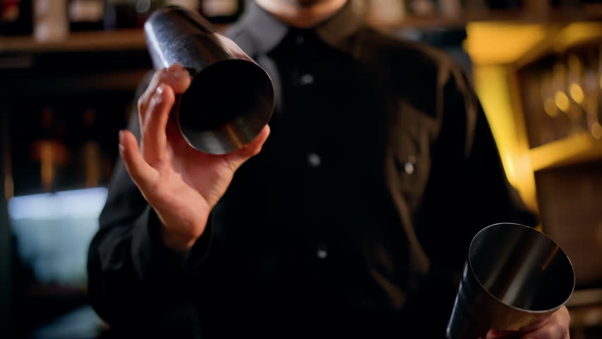 Close-up of a bartender in a dim bar assembling stainless steel cocktail shakers, preparing to mix a drink with precise hand movements amid warm ambient backbar lighting.