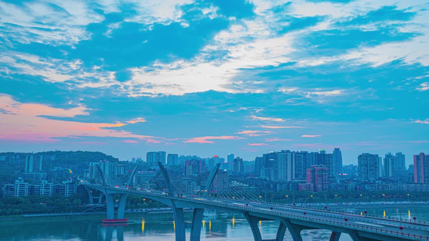 Modern cable-stayed bridge spanning river with urban skyline at dusk, dramatic cloudy sky with pink and blue hues creating stunning architectural cityscape view
