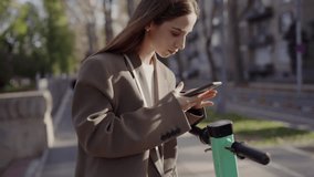 A young woman in a stylish coat uses her smartphone while standing beside an electric scooter on a busy city sidewalk. Young Woman Using Smartphone Near Electric Scooter in City - Powered by Shutterstock - Get 15% off with code: PIKWIZARD15