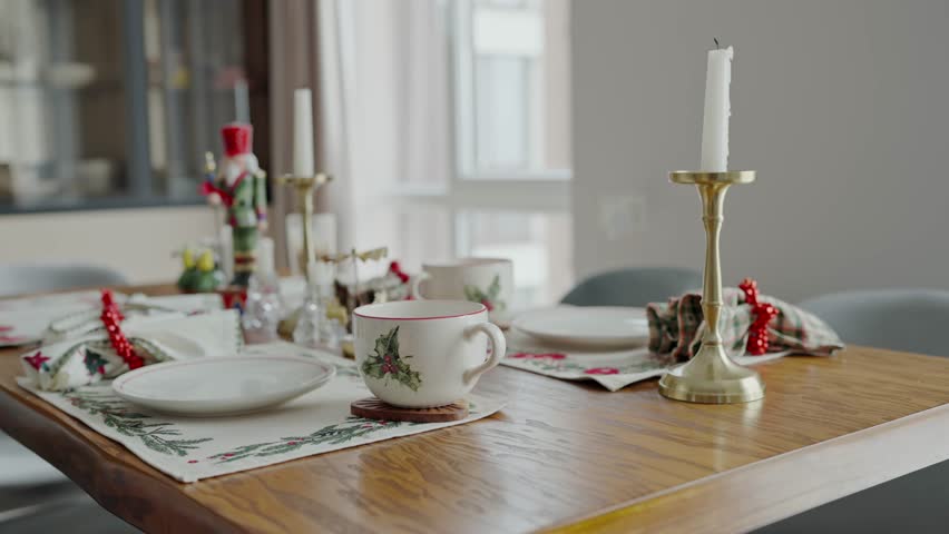 Festively decorated table setup for Christmas featuring a candle, holly themed ceramics, and holiday ornaments, capturing the essence of a warm, inviting Christmas dinner atmosphere.