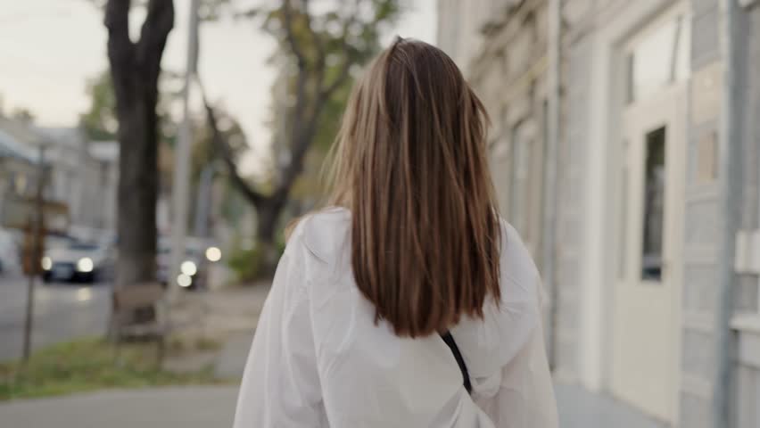 An image of a young woman, wearing a white blouse, smiling joyfully as she walks on a city street during a sunny day, adding a refreshing urban vibe. Young Woman Smiling as She Walks through a Street