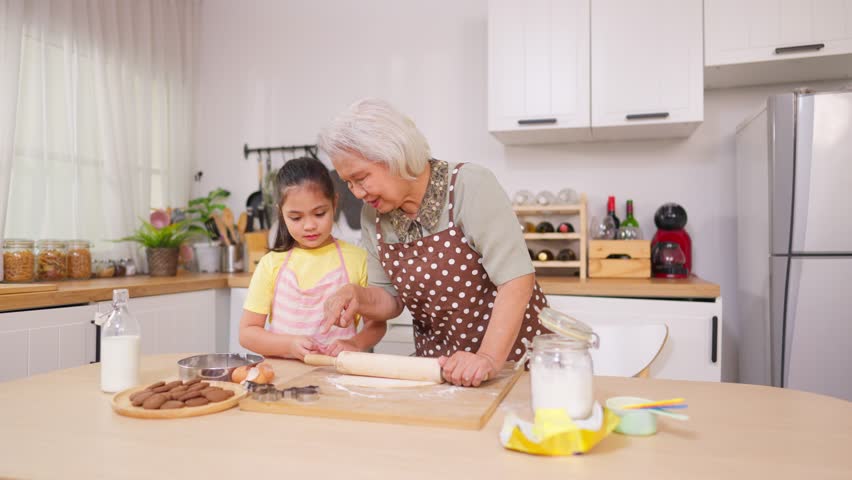 Asian granddaughter baking bakery with grandmother in kitchen at home. Happy family, adorable kid girl enjoys spending quality leisure time with caring senior mature grandma in cozy house together.