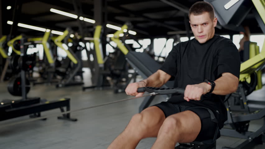 Blurred view of an athletic man working out in a modern gym, highlighting his focused approach and the gym's equipment. Young Man Exercising at a Modern Gym, Intense Workout Focus