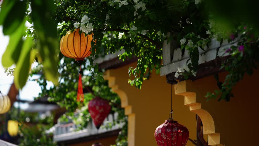 Close-up of traditional Vietnamese silk lanterns hanging from a yellow building in Hoi An, partially obscured by green leaves and white flowers.