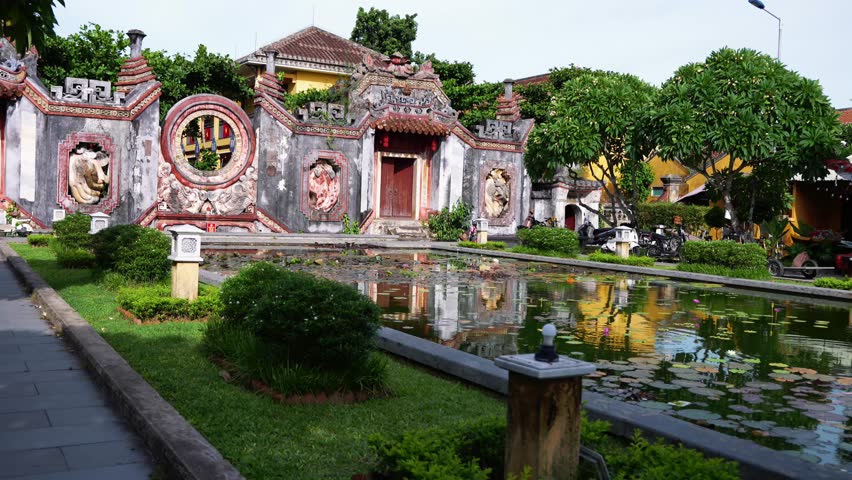 A beautifully weathered temple facade in Hoi An, Vietnam, features intricate carvings, a central doorway, and unique circular window, reflected in a tranquil foreground pond