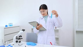 A young Asian female scientist holds a clipboard and a test tube filled with blue liquid, carefully reviewing experiment data related to science, laboratory, scientist, experiment, and research. - Powered by Shutterstock - Get 15% off with code: PIKWIZARD15