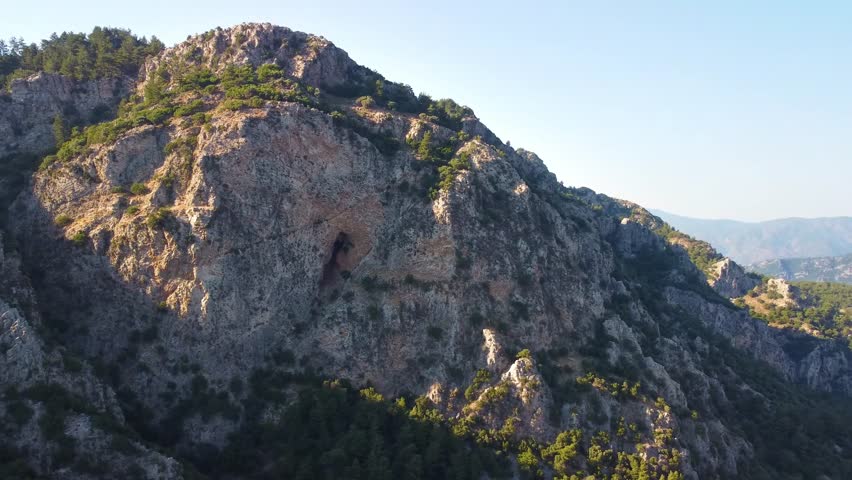 An aerial or high angle view showcasing a majestic and rugged mountain landscape featuring prominent rocky cliffs, textured rock formations, and scattered green vegetation under a clear sky. This natu