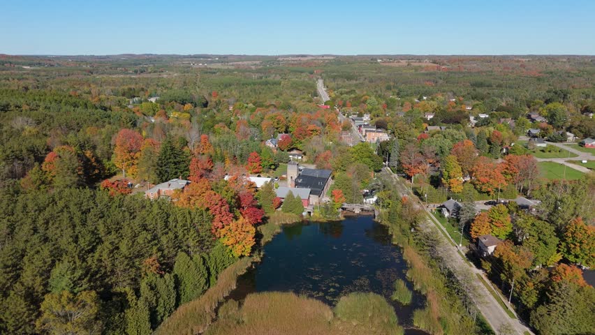 A beautiful drone shot of the historic Alton Mill Arts Centre, nestled beside a millpond and enveloped by a forest of vibrant fall foliage in rural Ontario, Canada.