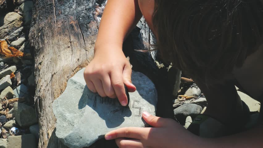A Child Writes With Natural Charcoal on a Stone Slow Mo. Seashore, nature and people.