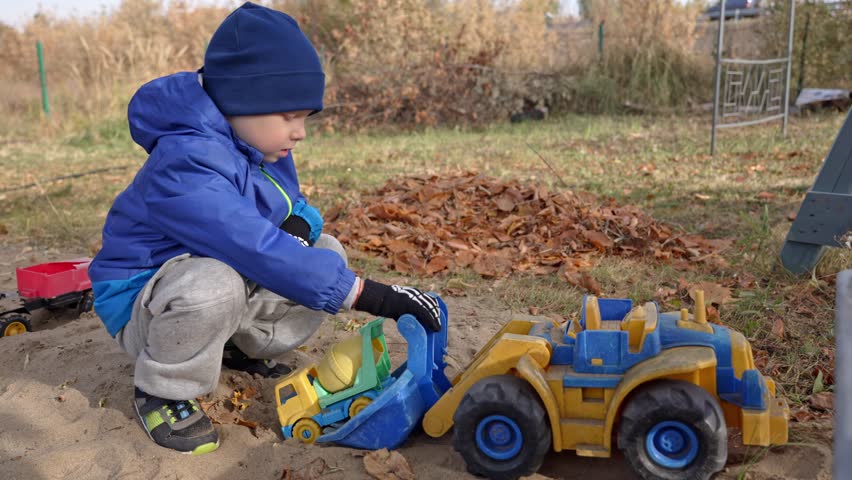 Young child wearing a blue jacket and hat engrossed in playing with a toy cement mixer and a large front loader truck in a sandy playground during a cool autumn day outdoors