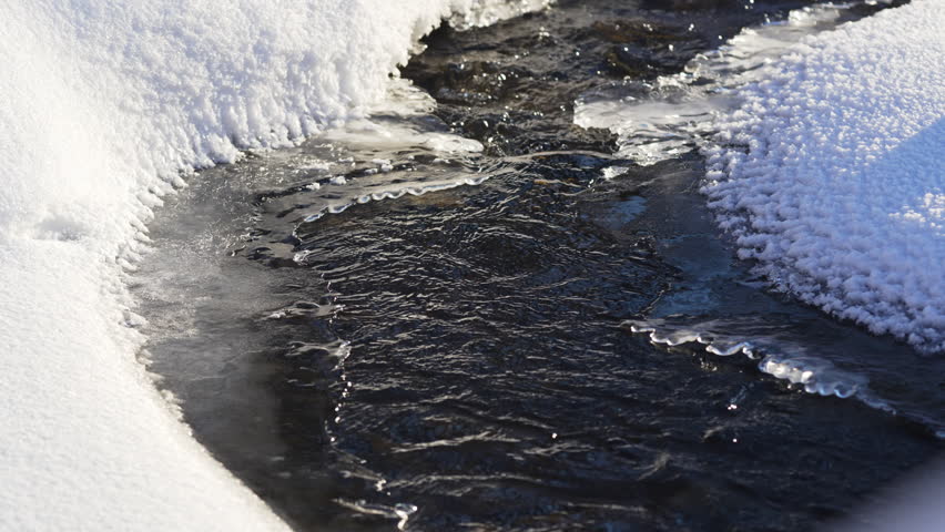 Flowing water glimmers between snowbanks forming mirrorlike path. Valley divided by shining stream shows perfect contrast of life and frost