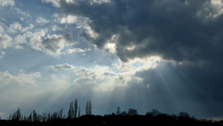Sunlight breaks through storm clouds creating shafts of light over landscape with trees. Bright light behind clouds contrasting dark silhouette.