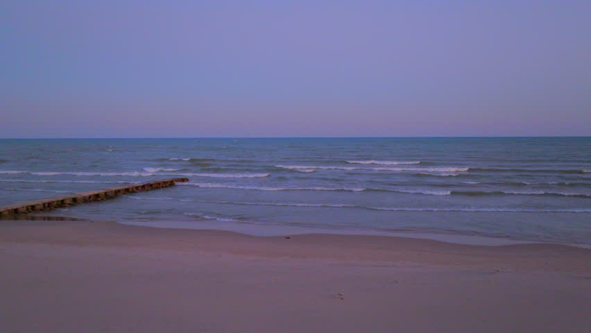 Drone view of gentle waves beside a concrete pier at a quiet, empty Lake Michigan beach in Sheboygan, Wisconsin, during dusk with a soft colorful sky on the horizon.