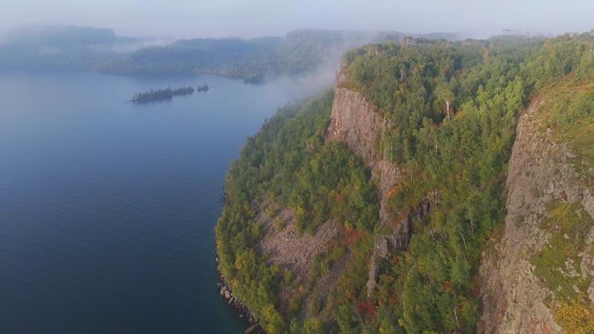 Sunlight illuminates Ontario’s wild Lake Superior shoreline, where bold cliffs rise above a forest glowing with early fall hues aerial view