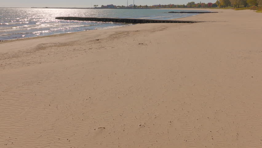 Drone aerial flying low from the beach over a concrete pier and Lake Michigan in Sheboygan, Wisconsin, as sunlight shimmers on gentle autumn waves on a clear, beautiful day.