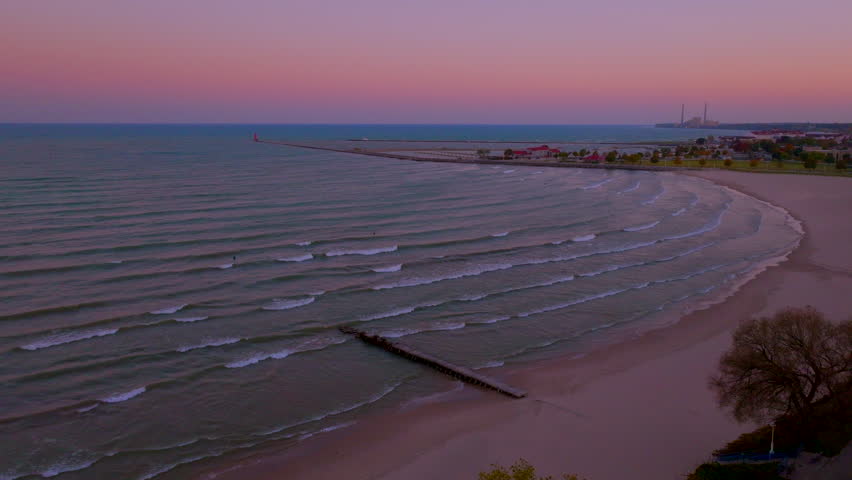 Drone aerial at dusk pushing toward a concrete pier stretching into Lake Michigan in Sheboygan, Wisconsin, under a soft pink and purple ombré sky with calm evening waves.