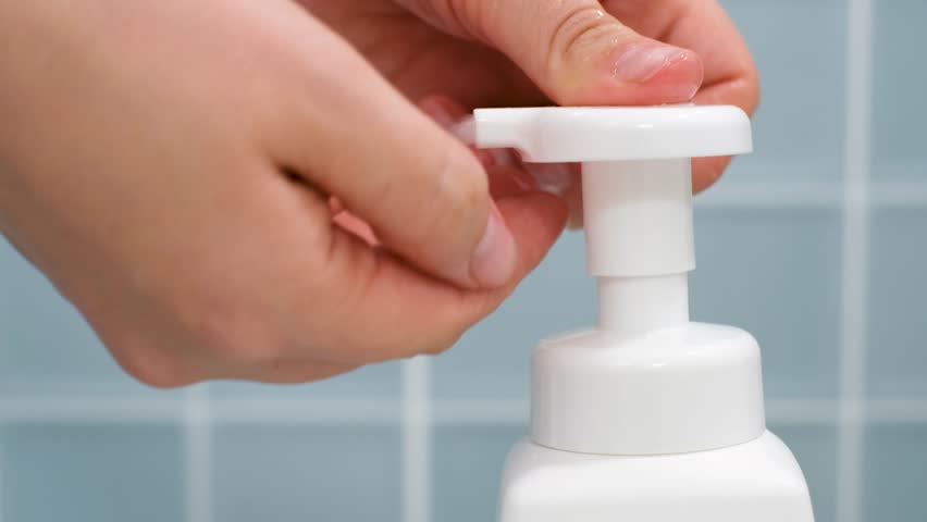 Hands squeeze liquid soap with foam from white bottle on blue tile background.