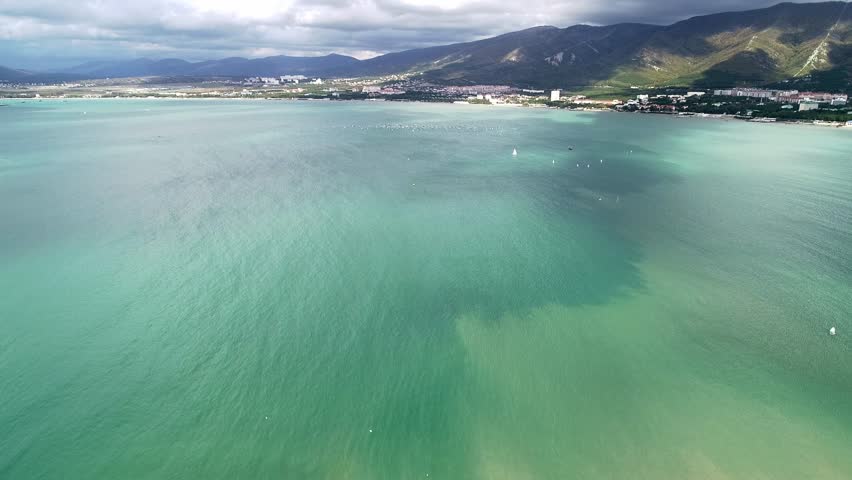 Yacht competitions in Gelendzhik Bay. Many small yachts on water. Aerial view. Caucasus Mountains in background. Clouds in sky. Green water of bay. Children's regatta