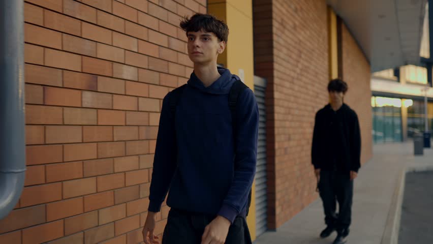 A young thug in black hoodie snatches a teenage boy is backpack on the street near a brick wall, showing urban crime, fear, and confrontation between two teens.