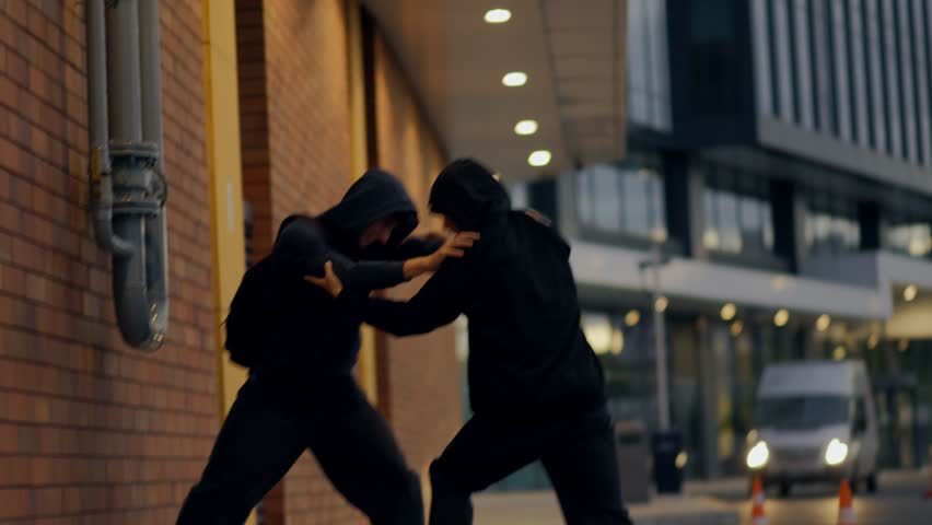 Two teenage boys wearing hoodies push each other in a violent street fight at night, reflecting crime, anger, fear, and urban social problems.