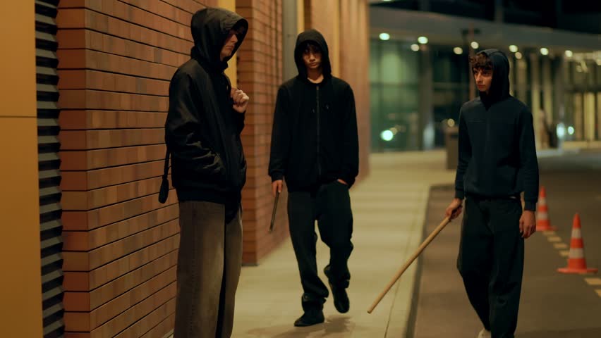 Two hooded teens menace another boy with sticks at night on the street. The image highlights crime, aggression, intimidation, fear, and serious urban problems.