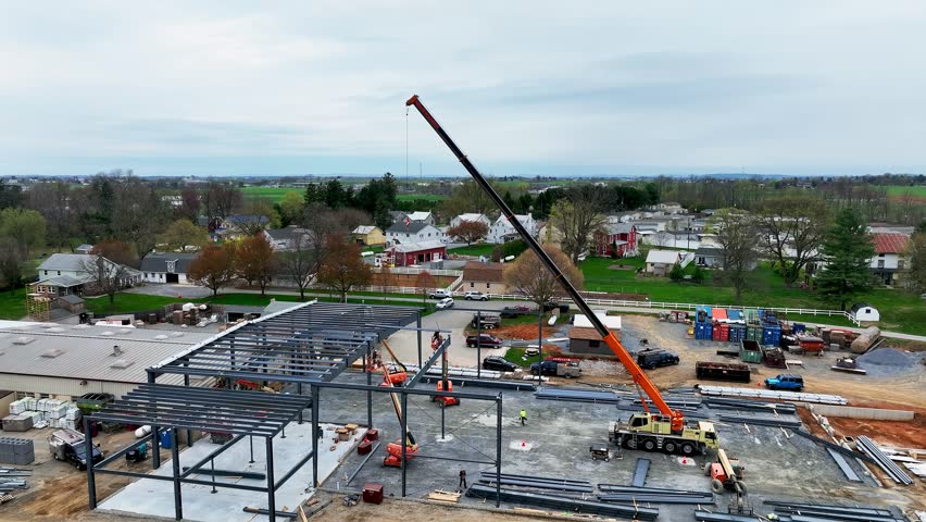 A construction site is busy with workers coordinating activities. A large crane is lifting materials to build a new structure. The setting is surrounded by houses and farmland on a cloudy day.