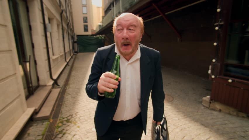 Wide angle close-up of a drunk man in a suit drinking alcohol outdoors. His reckless and inappropriate actions in public reveal hopelessness, sadness, and inner turmoil.