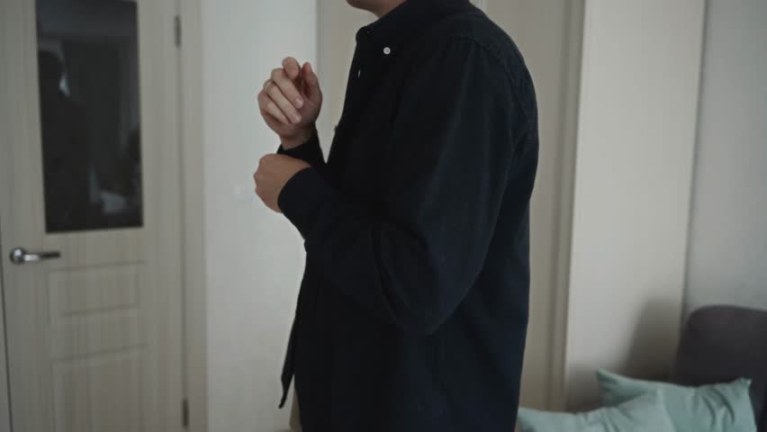 Medium shot of a man adjusting the cuff of his black shirt indoors. The frame reflects neat lifestyle, simple daily habits, and minimal home style. High quality 4k footage