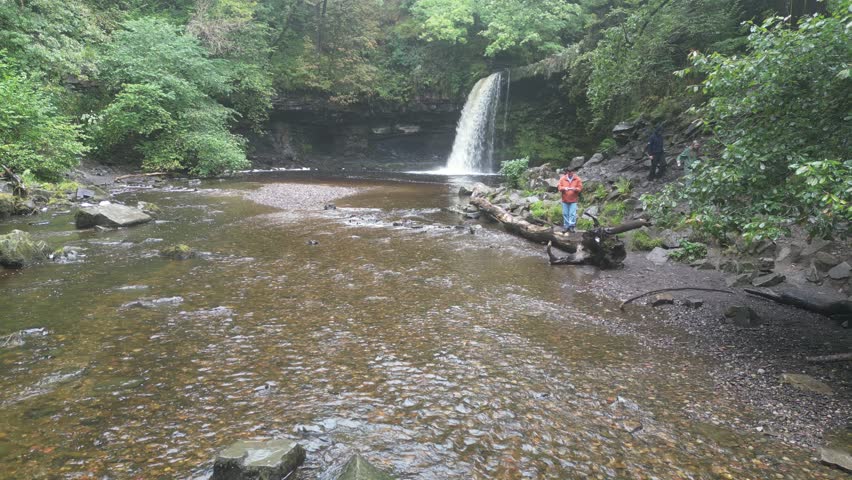 Aerial footage captures the serene Angel Falls waterfall in Brecon Beacons National Park, Wales. Wet, windy weather, lush foliage, and cascading water create a dramatic, natural scene.