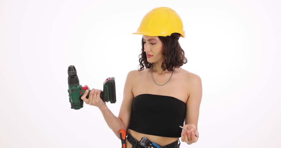 Woman builder in helmet looks at drill and steel bit expressing confusion. Female construction worker tries to figure out tool exploitation in studio