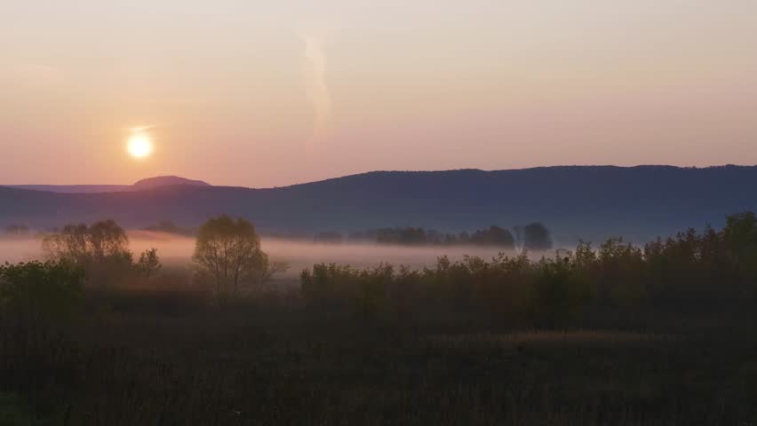 A serene and atmospheric footage capturing the golden sunrise over the misty South Ural mountains in the Republic of Bashkortostan, Russia. The morning fog blankets the valleys and trees, creating a t