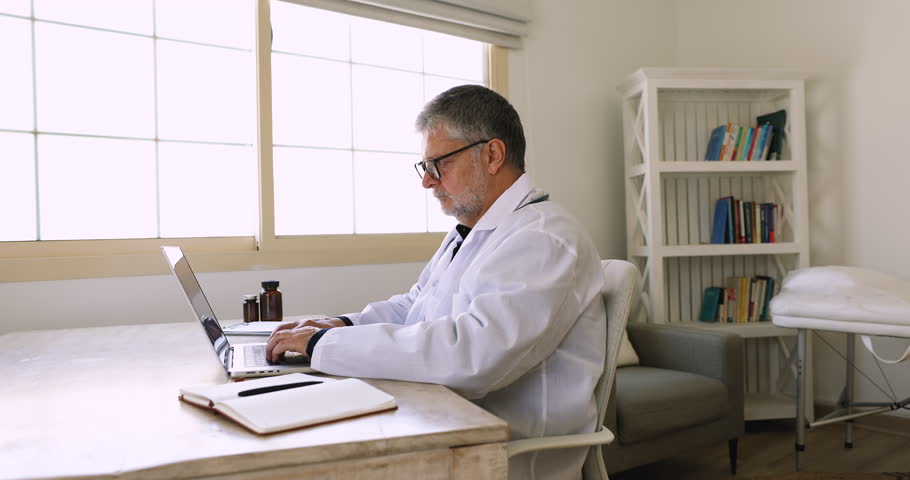 Older 55s Latino male doctor working online using laptop, provide professional medical consultation to clinic patients remotely, accessing medical card, writing prescription. Modern tech in healthcare