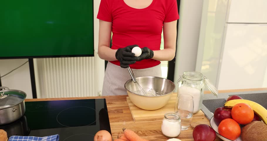 Woman in gloves cracks egg into metal bowl with dough mix in kitchen. Housewife cooks pastry of organic ingredients at wooden countertop with fruits