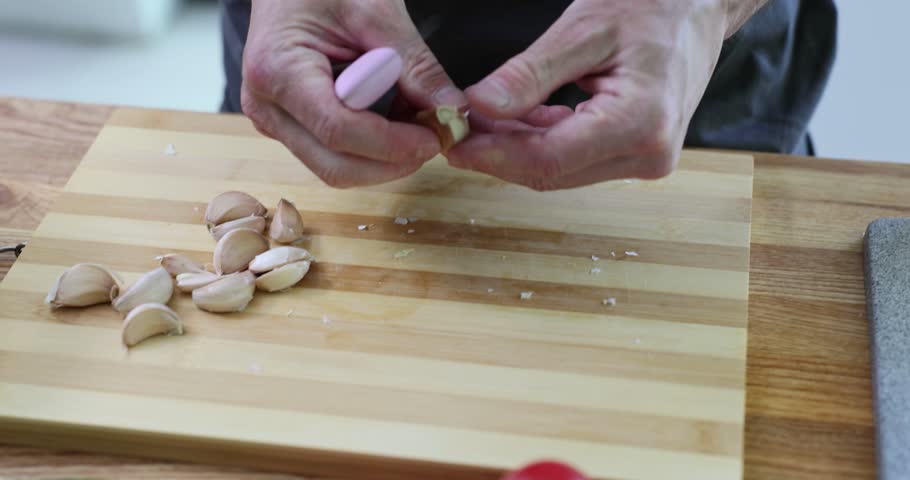 Man chef carefully peels garlic cloves using sharp knife at kitchen counter. Professional cook removes thin skin from garlic cloves. Slow motion