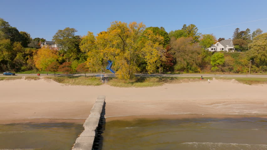 Drone aerial pulling back over Lake Michigan beach and gentle waves past a concrete pier, revealing shoreline road, houses, and autumn trees in Sheboygan, Wisconsin.